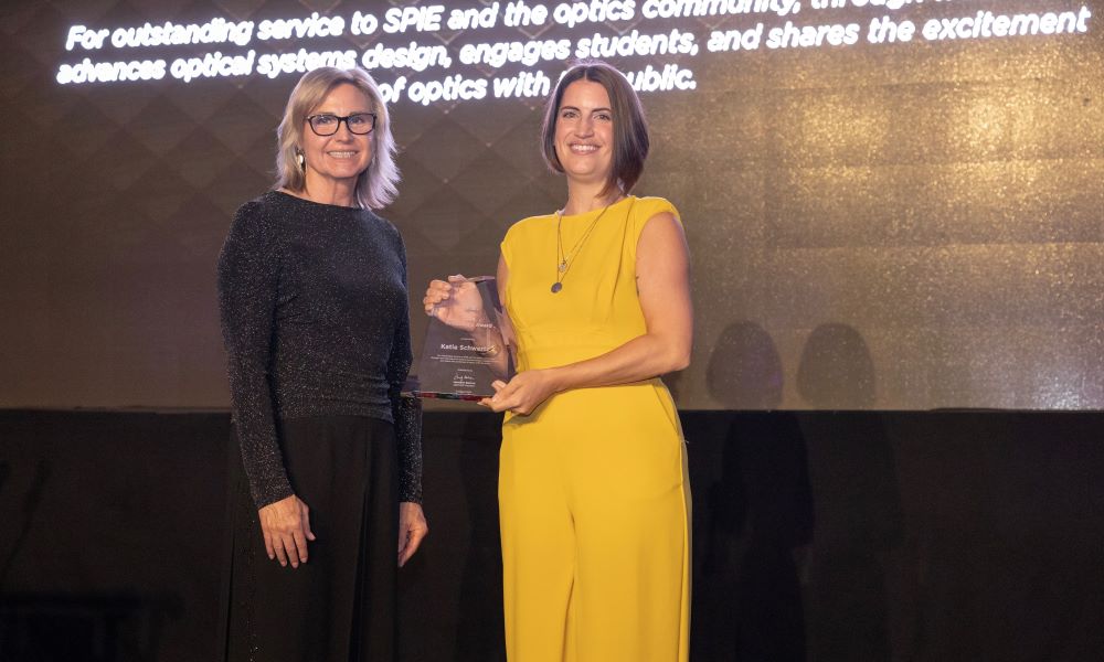 Katie Schwertz stands on the stage, holding her President's Award, with 2024 SPIE President Jennifer Barton