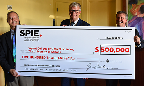 Three people holding up an oversize check for $500,000, made out to the Wyant College of Optical Sciences, The University of Arizona