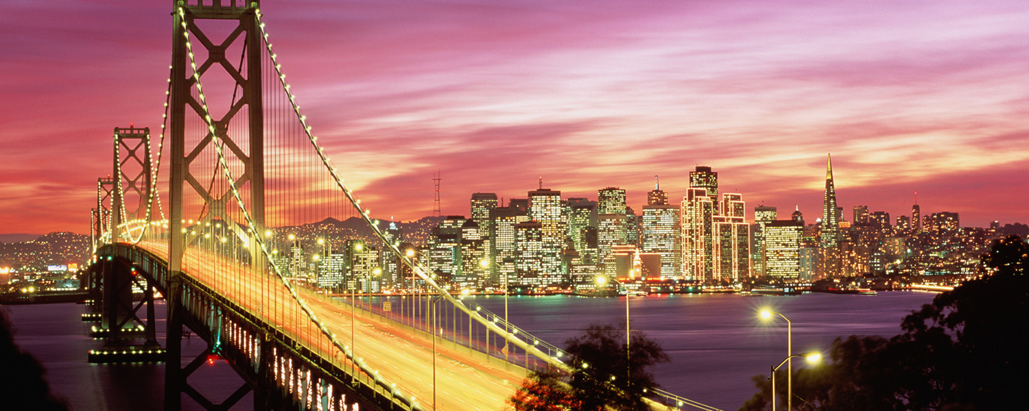 The Bay Bridge in the foreground with San Francisco city lights lining a colorful California sunset