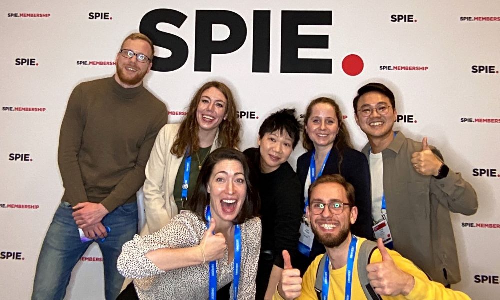 A group of seven smiling people with thumbs up, in front of the SPIE Membership photo booth backdrop