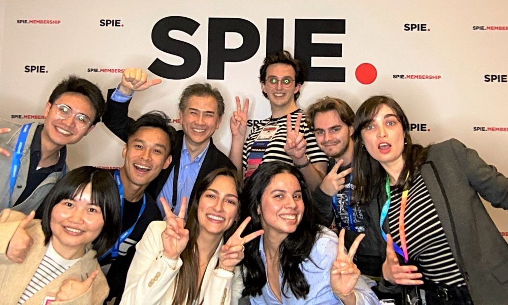 A group of nine smiling people posing in front of the SPIE Membership photo booth backdrop