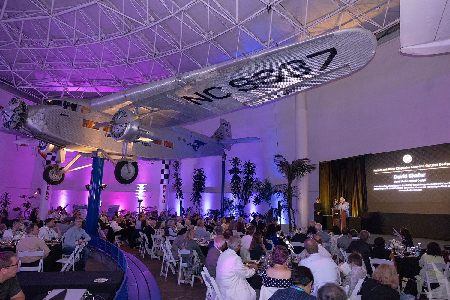 An airplane is perched overhead as the audience enjoys the award presentation of the  Rudolf and Hilda Kingslake Award in Optical Design to David Shafer at the 2024 Society Awards banquet.