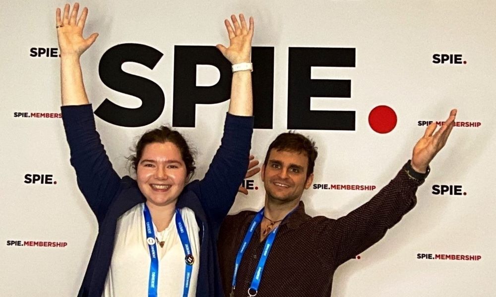 Two Student Members pose with their arms extended upward in front of the SPIE photo booth backdrop