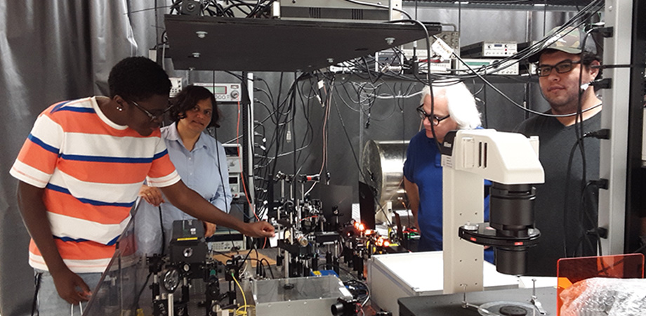 Experimental setup for a magnetometer using alkali sodium atoms in Tripathi's DSU lab. The yellow glow from a sodium laser is visible in the picture. Left to right: Lawrence Taylor, a freshman pursuing a BS in Engineering Physics; Tripathi; Robin Depto, a BS Electrical Engineering student at Delaware Technical Community College; and Mauricio Pulido who pursuing an MS in Applied Optics.